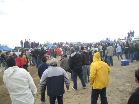 1990 Ford Bronco - large crowd in jackets lines a sandy off-road course with hills and blue canopy tents in the distance.