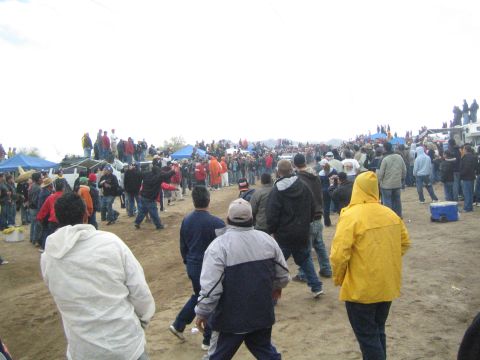 1990 Ford Bronco - Spectators gather on sandy terrain watching an off-road event, with a person in a yellow rain jacket and blue tents in the background.