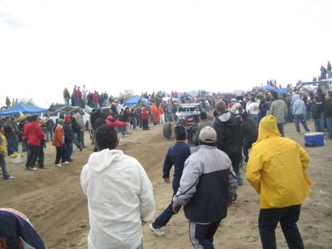 1990 Ford Bronco - crowd in jackets watches a tube-frame off-road buggy churn across a sandy course, with blue canopy tents and a cooler nearby.