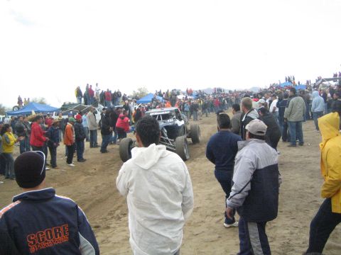 1990 Ford Bronco - white tube-frame off-road buggy navigates a sandy course amid a dense crowd, with a spectator in a SCORE jacket in the foreground.