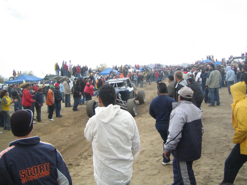 White off-road buggy on sand surrounded by crowd, man in SCORE jacket foreground