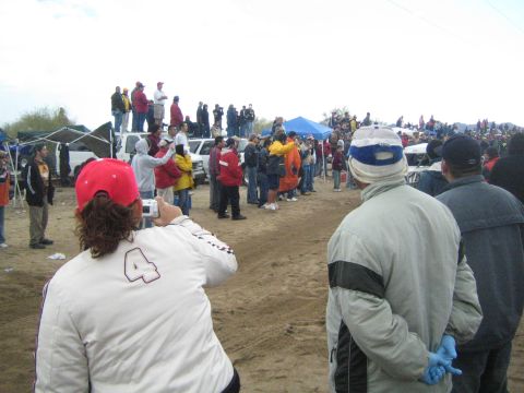 1990 Ford Bronco - Spectators line a dirt off-road course as a woman in a numbered jacket photographs the action near parked vehicles and tents.