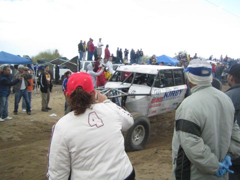 1990 Ford Bronco - white tube-frame off-road race buggy bearing Kirby and other sponsor decals sits on a sandy course as spectators watch.