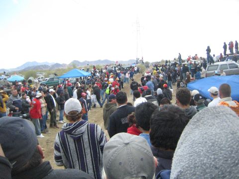 1990 Ford Bronco - dense crowd in jackets and caps lines a dirt off-road course, with a power transmission tower and distant hills behind.