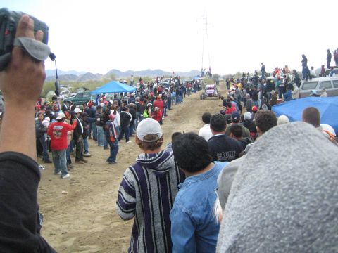 1990 Ford Bronco - spectator films a distant off-road buggy on a long sandy course flanked by crowds, tents, and a transmission tower.