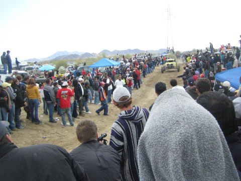 1990 Ford Bronco - Crowds in jackets line both sides of a sandy off-road course as a buggy approaches, with mountains in the background.