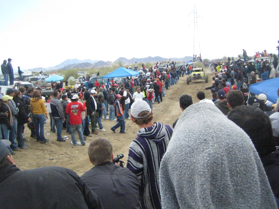 Crowds line a sandy off-road course with a buggy approaching and hills behind