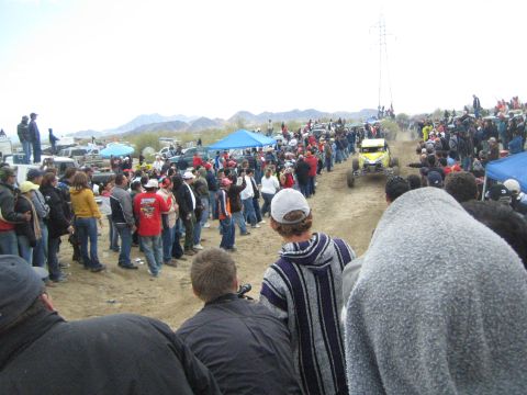 1990 Ford Bronco - yellow tube-frame off-road buggy speeds down a sandy course lined by large crowds, with desert hills behind.