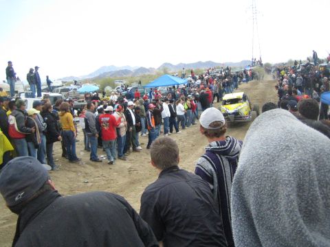 1990 Ford Bronco - yellow off-road buggy runs a sandy course as spectators in jackets watch, with a blue canopy tent and hills beyond.