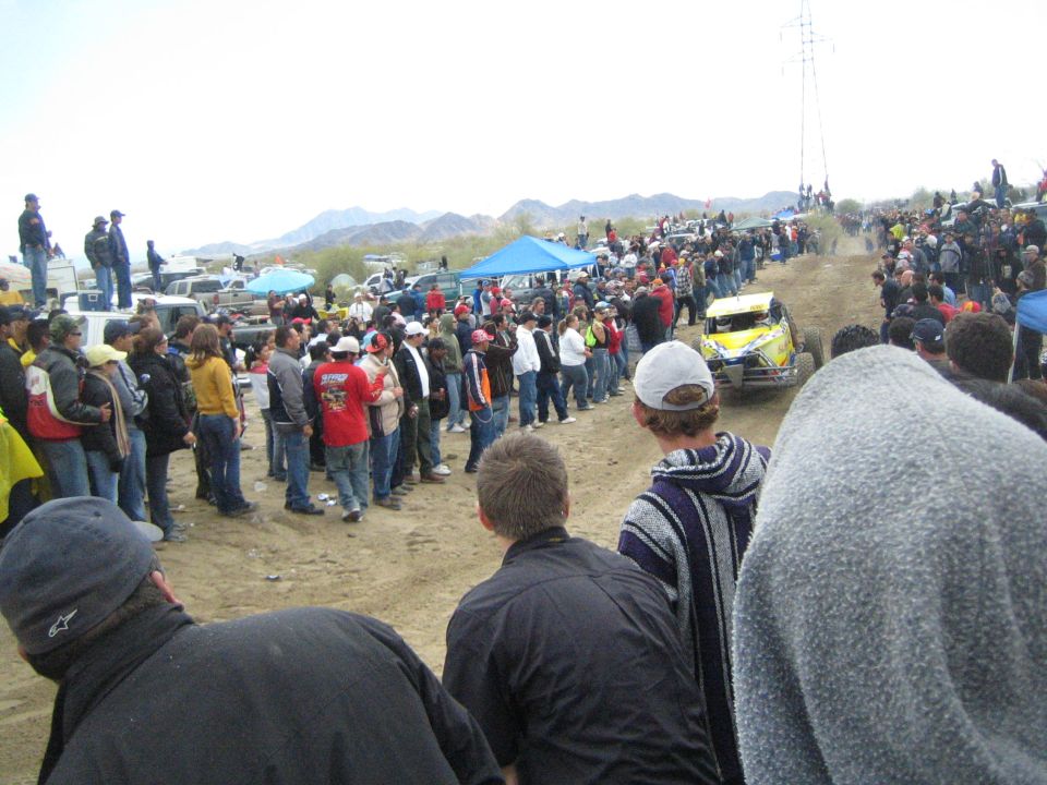 Yellow off-road buggy on sand with crowds and blue canopy tent in background