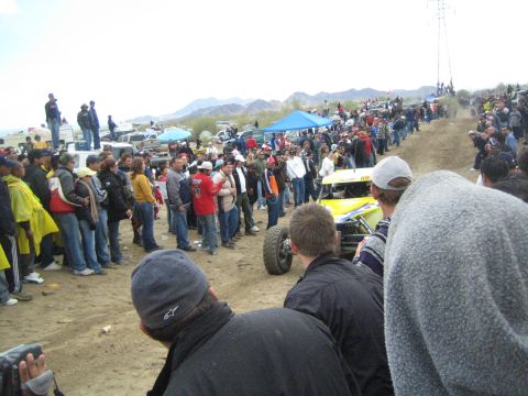 1990 Ford Bronco - yellow tube-frame off-road buggy kicks up sand on a course lined by spectators in cold-weather gear, mountains behind.