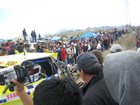 1990 Ford Bronco - spectator films a yellow off-road buggy marked 1010 as it sits on a sandy course amid a large crowd.