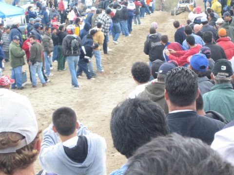 1990 Ford Bronco - Spectators in jackets and caps watch from a sandy berm as people walk along a dirt off-road course.