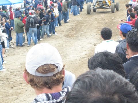 1990 Ford Bronco - yellow tube-frame off-road buggy runs a sandy course flanked by dense crowds of spectators in jackets and caps.
