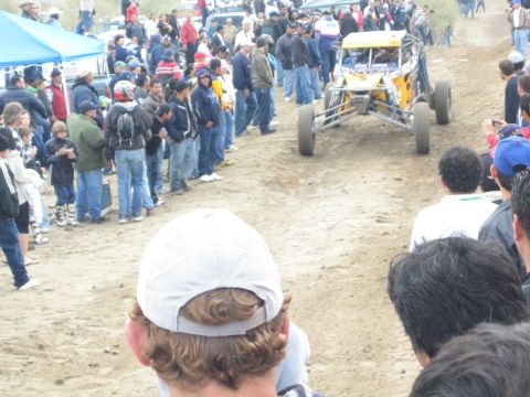 1990 Ford Bronco - yellow off-road buggy navigates a sandy course as spectators line both sides, with a blue canopy tent in the background.