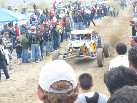 1990 Ford Bronco - yellow and white tube-frame off-road buggy crawls a sandy course amid spectators in jackets, with tents nearby.