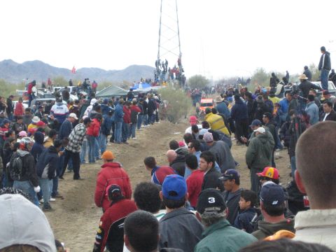 1990 Ford Bronco - large crowd lines a desert off-road course, with several people climbing a power transmission tower for a better view.
