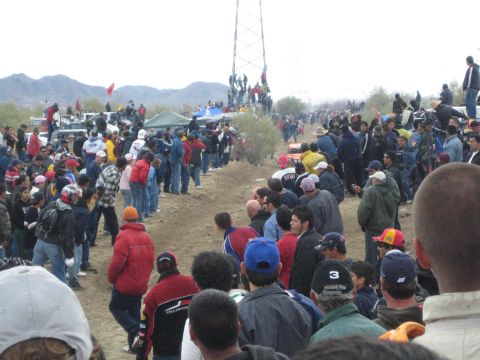 1990 Ford Bronco - massive crowd in jackets lines a desert dirt course, with spectators perched on a power transmission tower and hills beyond.