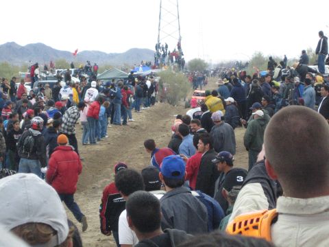 1990 Ford Bronco - dense crowd in cold-weather jackets fills a desert off-road course as spectators climb a power transmission tower in the distance.