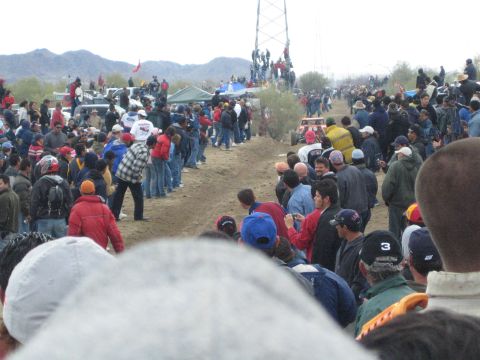 1990 Ford Bronco - Spectators pack both sides of a dirt desert race course, watching from behind as a distant buggy approaches under overcast skies.