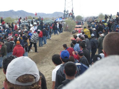 1990 Ford Bronco - small open-wheel off-road buggy runs the dirt course as a large crowd lines the track in front of low desert mountains.