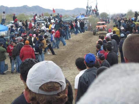 1990 Ford Bronco - Crowds line a winding dirt race course in the desert, watching an open-wheel buggy with people perched on a tower in the distance.