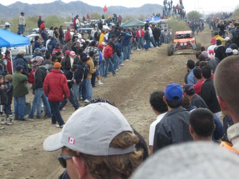1990 Ford Bronco - orange off-road buggy runs a dirt desert course lined with spectators; a foreground cap shows a Canadian flag.