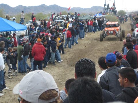 1990 Ford Bronco - orange off-road race buggy speeds along the dirt course as spectators and a blue canopy tent line the edge.