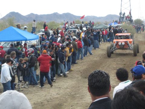 1990 Ford Bronco - orange open-wheel off-road buggy runs the dirt course past a dense crowd and a blue canopy tent in the desert.