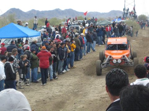 1990 Ford Bronco - orange off-road buggy marked BFGoodrich and number 1000 runs the dirt course past spectators and a blue tent.