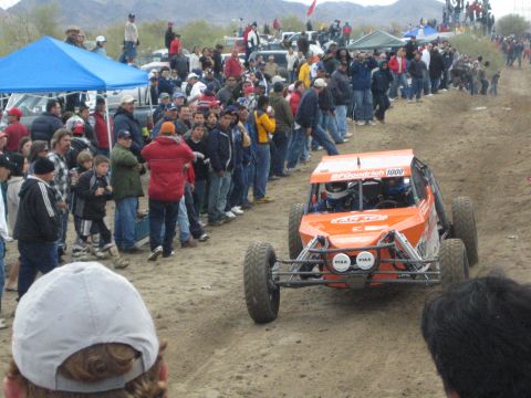 1990 Ford Bronco - orange off-road buggy bearing BFGoodrich and PIAA decals and number 1000 drives the dirt course past a crowd.