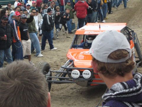 1990 Ford Bronco - helmeted driver pilots an orange off-road buggy with BFGoodrich branding and twin round PIAA lights past close spectators.