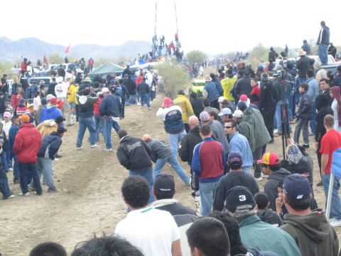 1990 Ford Bronco - Spectators gather and cross a dirt desert race course, with people watching from a tall tower structure in the distance.