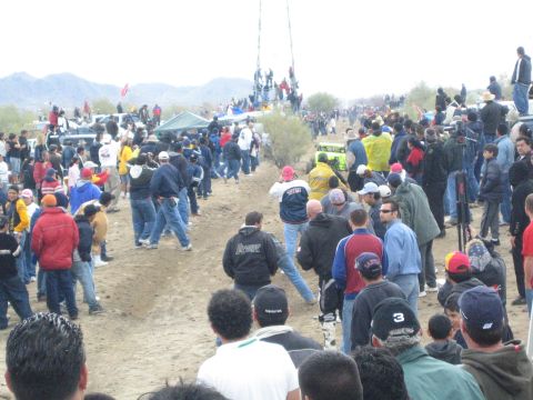 1990 Ford Bronco - large crowd fills a dirt desert course as a small off-road buggy is visible in the distance under gray skies.