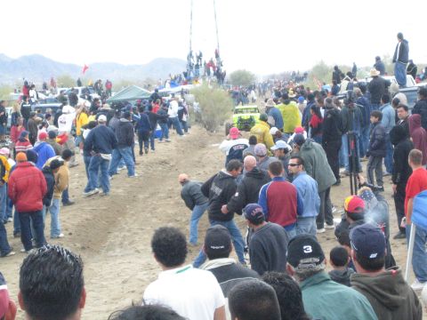 1990 Ford Bronco - Crowds line and cross a dirt desert race course while a distant off-road buggy runs the track near low mountains.