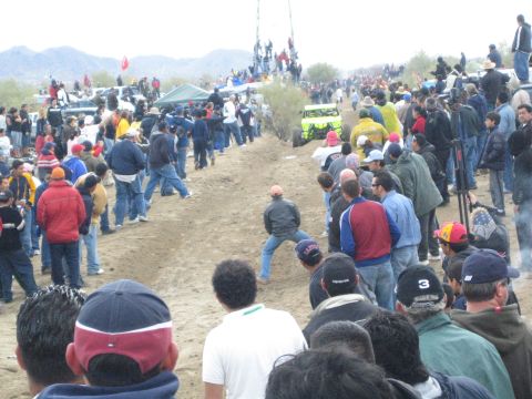 1990 Ford Bronco - green off-road buggy runs the far dirt course as dense crowds line the track in the desert under overcast skies.