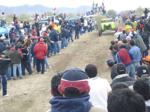 1990 Ford Bronco - yellow-green off-road buggy drives the dirt course past spectators lining the desert track near low mountains.