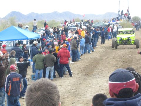 1990 Ford Bronco - bright green off-road buggy drives the dirt desert course past a crowd and blue canopy tents.