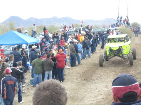1990 Ford Bronco - green-and-white off-road buggy runs the dirt course as spectators line the desert track under gray skies.