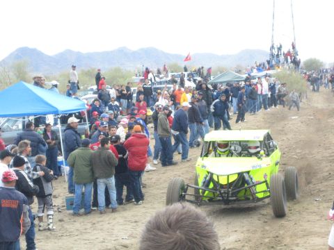 1990 Ford Bronco - bright green open-wheel off-road buggy approaches on the dirt course as a large crowd watches from the trackside.