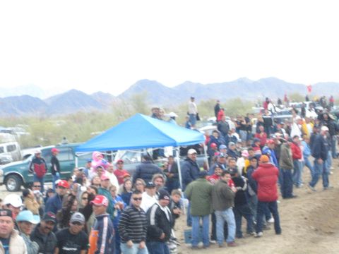 1990 Ford Bronco - Spectators gather around a blue canopy tent and vehicles in a desert race pit area beneath low mountains.