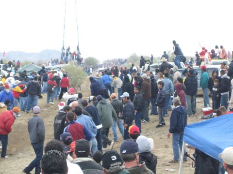 1990 Ford Bronco - dense crowd stands and mills on a dirt desert race course lined with parked trucks and canopy tents.