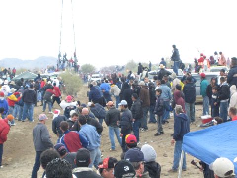 1990 Ford Bronco - Crowds cross and gather on a dirt desert race course with parked trucks and a viewing tower in the distance.