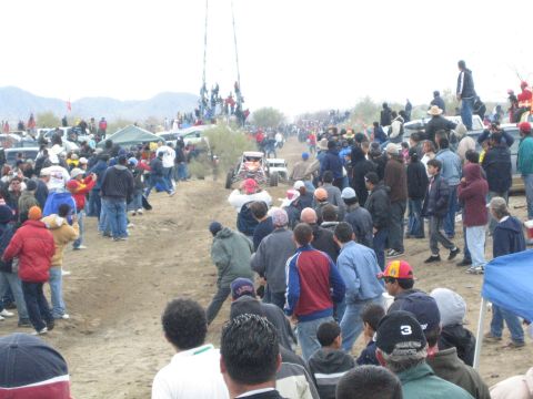 1990 Ford Bronco - dense crowd lines a dirt desert race course as off-road buggies run the track near a distant viewing tower.