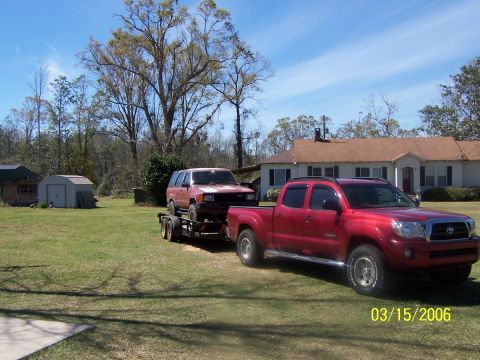 1984 Toyota 4Runner - A red pickup truck tows a red SUV on a trailer in front of a house.
