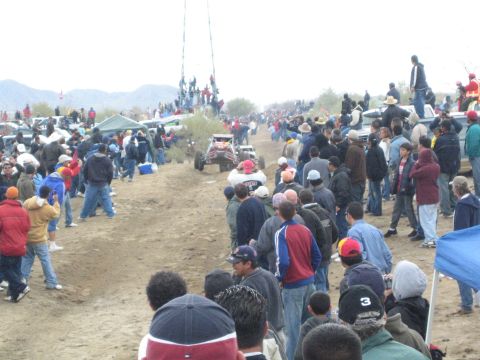 1990 Ford Bronco - single-seat off-road racing buggy rounds a dirt desert course as a dense crowd of spectators lines both sides of the track.