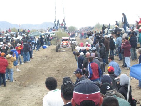 1990 Ford Bronco - open-frame off-road buggy climbs a rutted dirt track in the desert while crowds of onlookers watch from the edges.