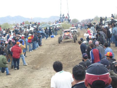 1990 Ford Bronco - red and white open-wheel off-road buggy speeds along a dirt desert course bordered by a thick line of spectators.