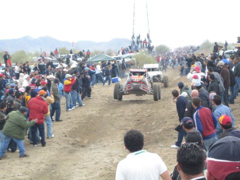 1990 Ford Bronco - off-road racing buggy drives a desert dirt course with another race vehicle ahead, surrounded by spectator crowds.