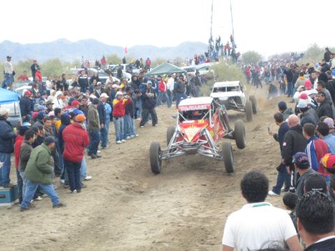 1990 Ford Bronco - red, flame-painted open-frame off-road buggy faces the camera on a dusty desert course as spectators line the track.